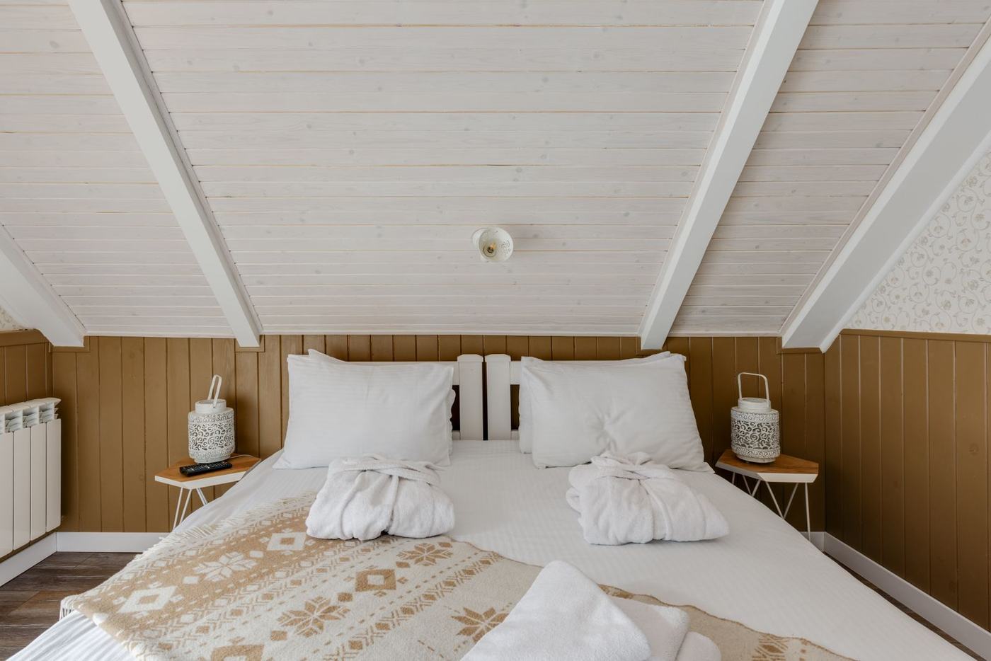 A cosy attic bedroom with white boarded ceiling, robes laid on the bed and lanterns either side
